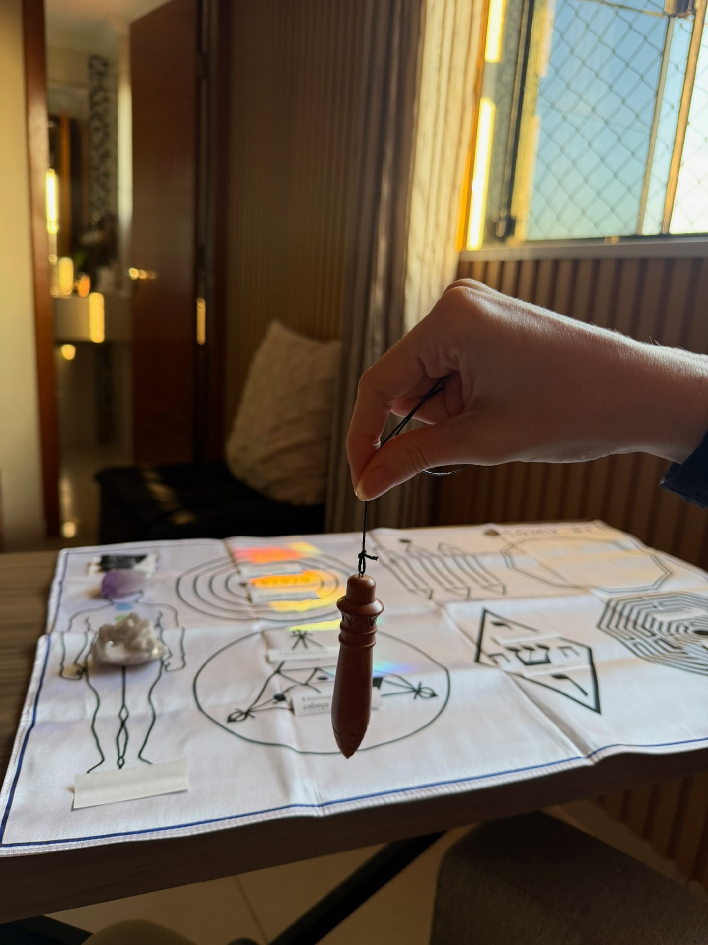 Mão segurando um pêndulo de madeira sobre uma mesa radiestésica