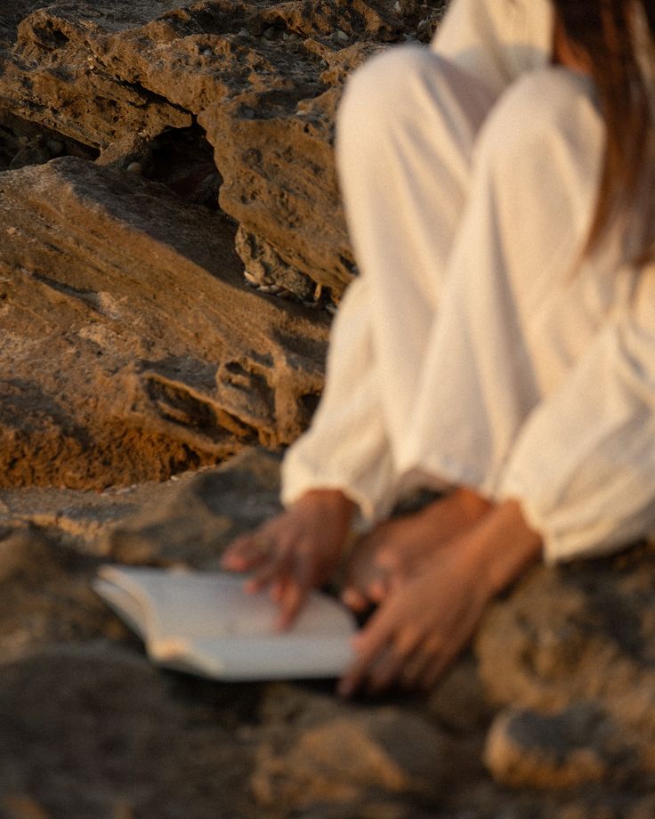Mulher sentada sobre pedras ao pôr do sol, com as mãos apoiadas em um caderno aberto